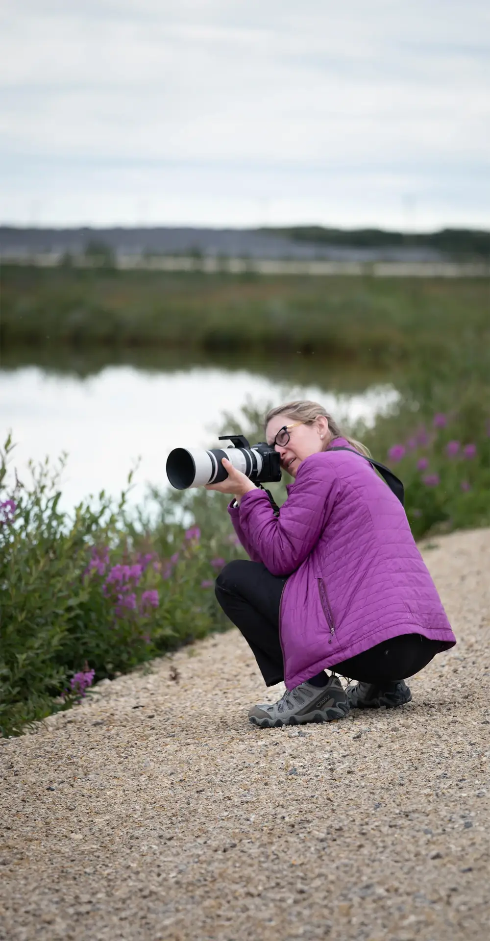 Traveler crouching with a telephoto lens beside a tundra pond and wildflowers