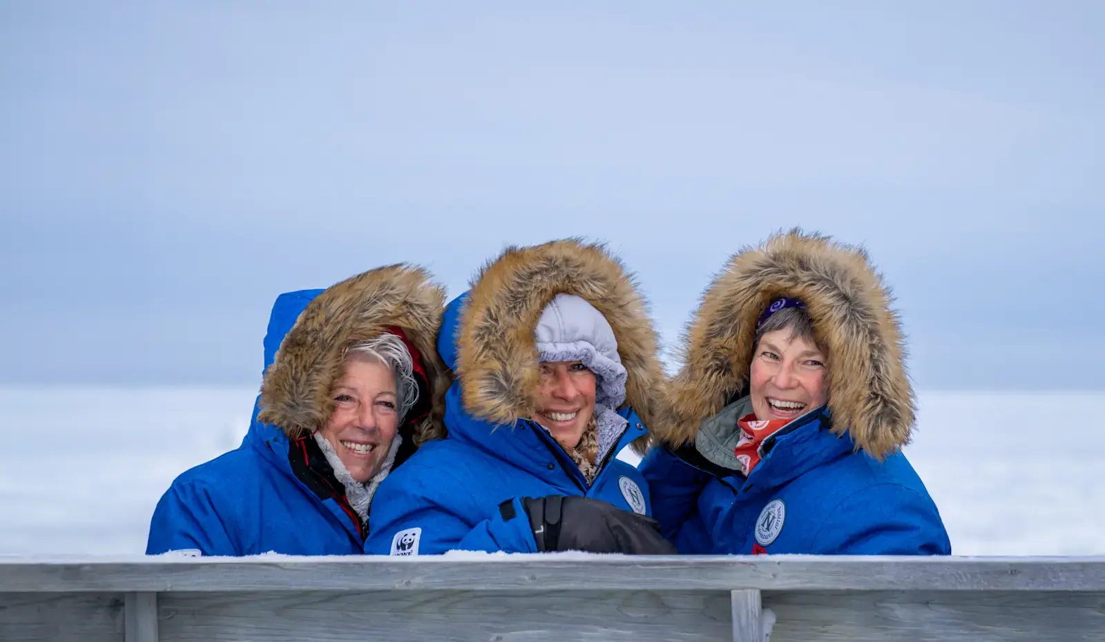 Three travelers in parkas smiling on a viewing deck above Hudson Bay